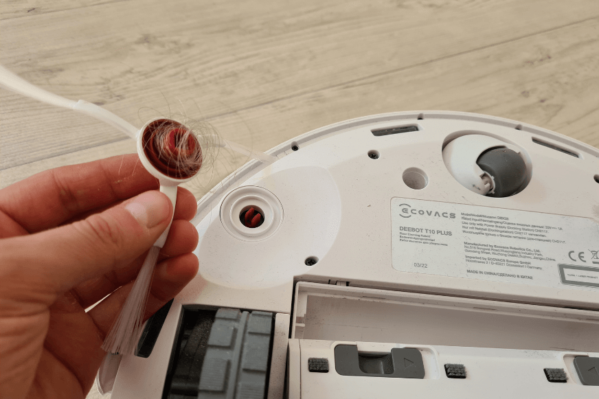 Die Seitenbürste des ECOVACS DEEBOT T10 wird abgenommen und die sich dort aufgewickelten Haare werden mit der Hand entfernt.